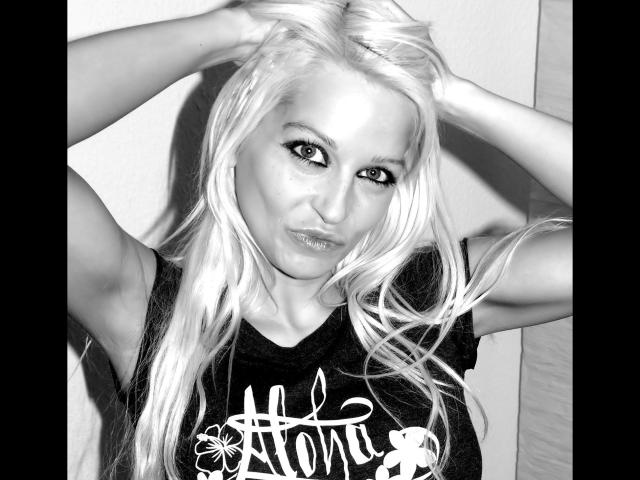 Black and white photo of jackylafey-hot with long platinum blonde hair, hands in hair, pouty lips, wearing a black t-shirt, looking directly at camera with sultry eyes.