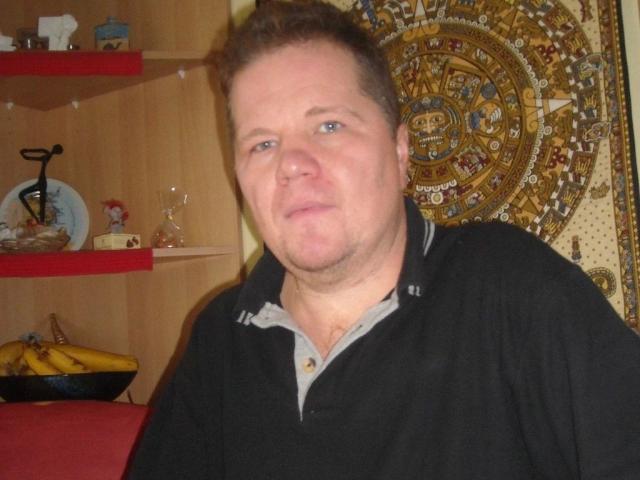 SPIKI-hot, chubby, middle-aged dude with short brown hair, wearing a black polo, gray shirt underneath, looking slightly annoyed, in a kitchen with wooden shelves and banana bowl.