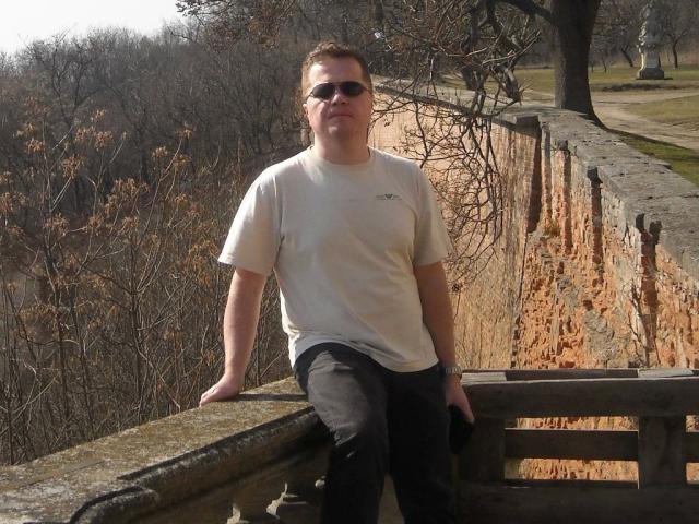SPIKI-hot with short brown hair, wearing sunglasses, white t-shirt, and black pants, leans on a stone wall, arms relaxed, looking slightly to the side, in daylight.