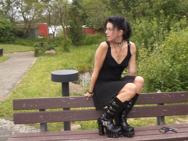 Carosinder-hot with black hair, glasses, black dress, and studded boots sits on a bench, legs crossed, showing her black boots and black panties. Park setting, overcast.
