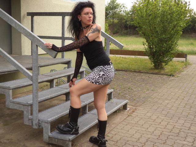 Carosinder-hot with wild black hair, black fishnet sleeves, black top, leopard print skirt, black boots, standing on metal stairs, showing her ass, tattoo on right arm, looking angry.