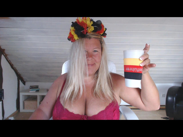 Kim-Van-Dyke with blonde hair, red lace bra, and flower crown, holding a beer mug, showing large boobs, smiling, in a white room.
