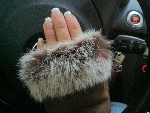Kim-Van-Dyke's hand with white nails, wearing a brown fur cuff, resting on a black car steering wheel. Keychain with red button visible.