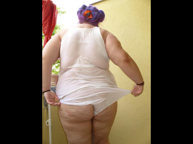 scharfe-kitty, rechoncha con piel pálida, vistiendo una camiseta blanca, mojada y sin mangas, levantándola para mostrar su trasero, cabello morado recogido en un moño, de pie frente a una pared amarilla..