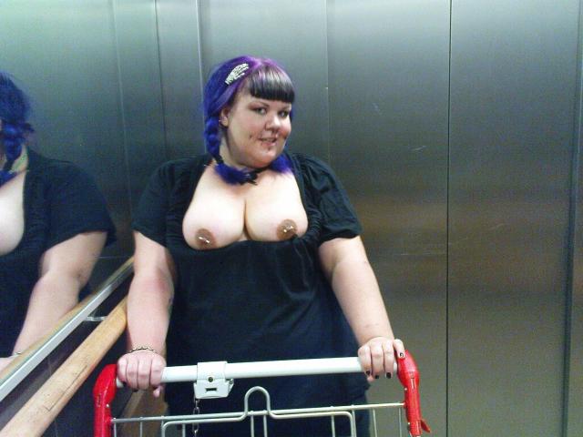 Scharfe-kitty mit lila Haaren, schwarzem Kleid und freiliegenden Brüsten, steht in einem Aufzug, hält einen roten, metallenen Rollstuhl, lächelt, Spiegelbild ist sichtbar..