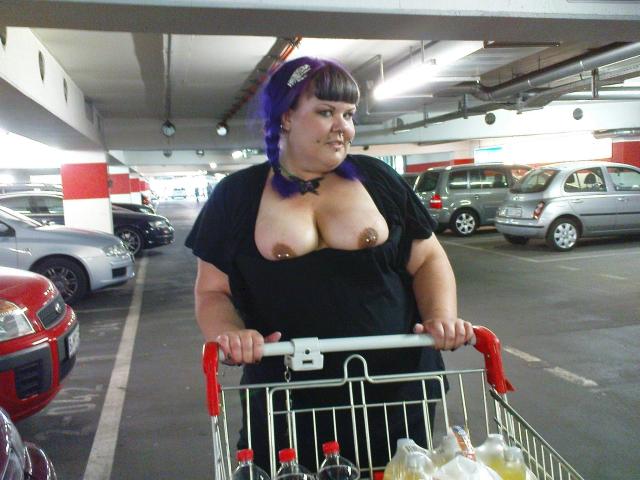 Scharfe-kitty con i capelli viola, una maglietta nera che mette in mostra un seno abbondante, che spinge un carrello della spesa in un parcheggio sotterraneo, tenendo le maniglie del carrello, che guarda di lato con un leggero sorriso..