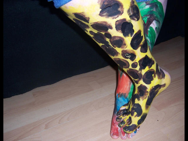 As pernas de scharfe-kitty, pintadas com manchas de leopardo em amarelo e preto vibrantes, e padrões coloridos em verde, azul e vermelho, estão cruzadas em um piso de madeira, mostrando dedos detalhados e pintados..