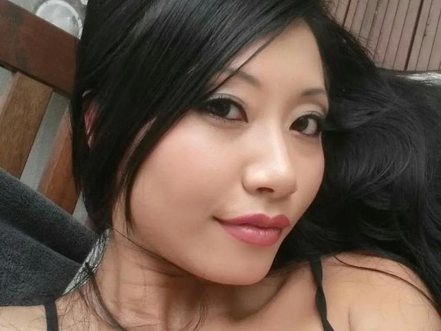 El rostro de Lady-Tyra, con piel asiática lisa, cabello negro y lápiz labial rosa, presenta un delineado de ojos sutil y una ligera sonrisa. Lleva una correa negra, y sus senos no son visibles..