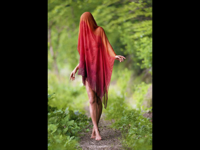 Lulu-4-you com um capuz vermelho-alaranjado cobrindo sua cabeça, caminhando descalça em uma trilha de terra, com os braços levemente levantados, em um ambiente verde, semelhante a uma floresta..