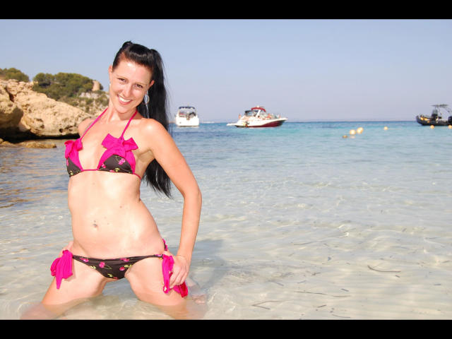 Texaspatti, con un bikini floral rosa y negro, de pie en agua clara, sonriendo, con cabello negro largo, pechos pequeños y un ombligo lindo, en una playa soleada..