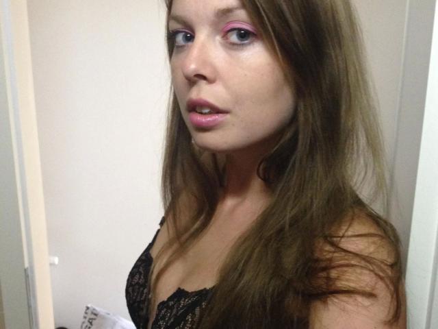 SeiMeinVoyeur-hot with long brown hair, pink eyeshadow, and black lace bra, showing her tits, pink lips slightly parted, standing in a room with a white door.