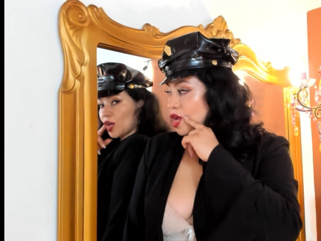 AlisGourmande with black wavy hair, wearing a black leather cap and black jacket, touches her red-lipped lips, showing her white bra-covered boobs in a gold-framed mirror.