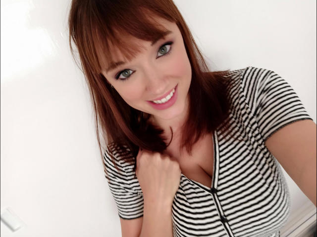 FannyXX-hot with brown hair and green eyes, smiling at the camera. She's wearing a striped top with a zipper, showing a little cleavage. Her hand is near her chest.