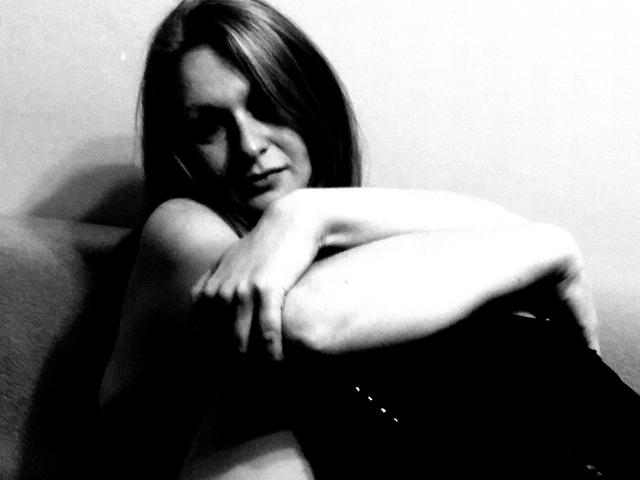 Black-and-white photo of SweetElisa1-hot with long hair, arms crossed over bare chest, looking slightly down, wearing black clothing, sitting against a light wall.