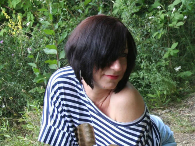 Inga-Star con i capelli neri corti, una camicia a righe, con una spalla scoperta, sorridente, con un leggero rossetto rosso, seduta all'aperto, in un'area erbosa, con piante verdi e piccoli fiori gialli, che tiene in mano un bastoncino marrone..