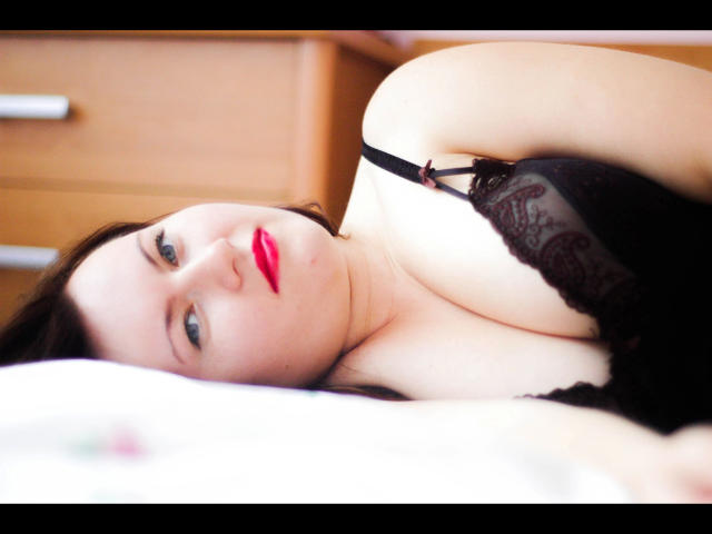 Fraumia, con il rossetto rosso, sdraiata su un letto, indossa biancheria intima di pizzo nero, mostrando il suo seno prosperoso e la sua pelle chiara, guarda verso l'alto con un'espressione seducente..