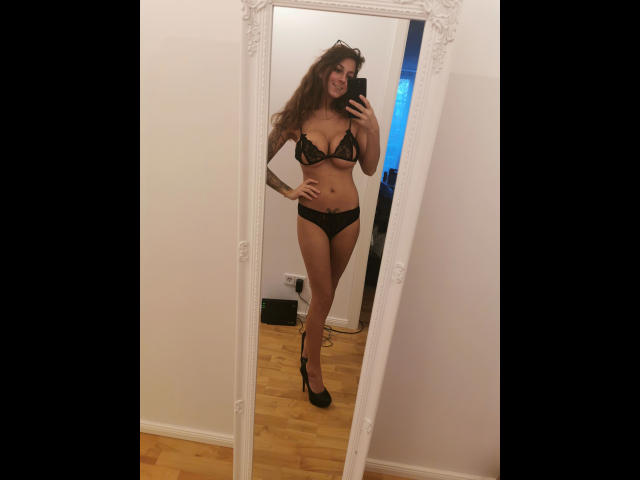 Autoportrait de KaitySun, magnifique en lingerie noire en dentelle, montrant sa poitrine et ses fesses, avec de longs cheveux bruns, un tatouage sur le bras gauche, et des talons hauts noirs, debout sur un plancher en bois..