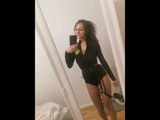 KaitySun, vêtue d'une robe noire à manches longues, à fermeture éclair, taille haute, avec jarretières et courte, sourit devant un miroir, tenant un téléphone, avec des cheveux bruns bouclés et une posture confiante..