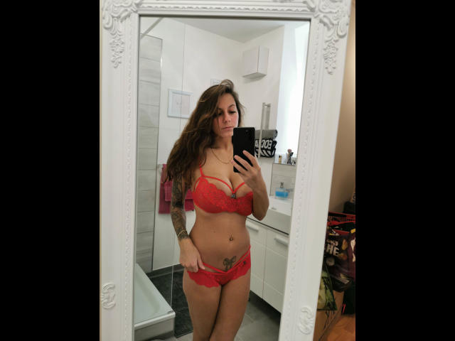 KaitySun-hot se tient devant un miroir, vêtue de lingerie en dentelle rouge, montrant ses seins et un tatouage sur sa hanche droite, tenant un téléphone, avec de longs cheveux bruns et un sourire confiant..