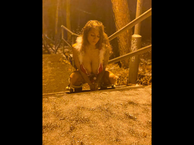 KaitySun sitzt in einem roten Top mit Fellbesatz, das ihre großen Brüste zeigt, auf einem schmutzigen Pfad, mit lockigem Haar und einem verführerischen Ausdruck, in einem von goldenem Licht durchfluteten Wald..