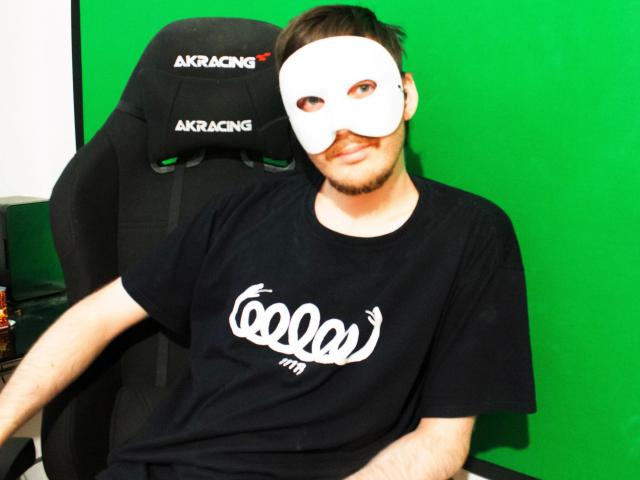 Sweetbody23-hot wearing a white mask, black t-shirt with white logo, and black chair, sitting with green background, has short dark hair and a slight beard.