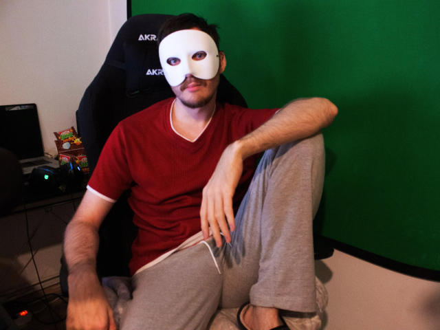 Sweetbody23-hot with a white mask covering his face, wearing a red shirt, gray pants, and sandals, sitting in a black chair with a green background.