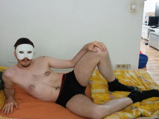 Sweetbody23-hot with a white mask, hairy chest, black underwear, and black socks, laying on a yellow bedspread, legs spread, showing a flaccid, uncircumcised penis.