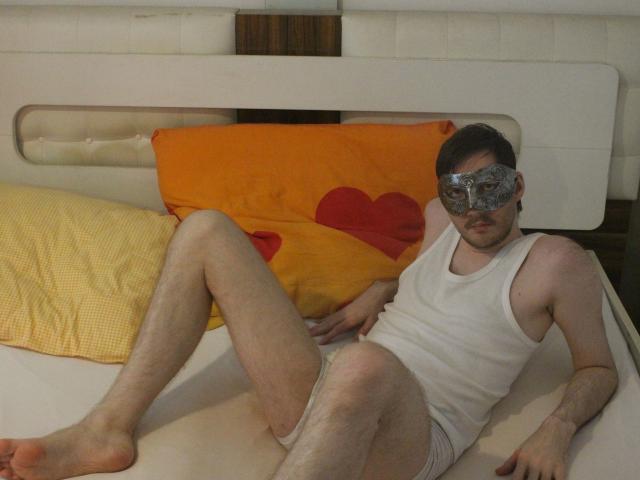 Sweetbody23-hot, wearing a white tank top and gray underwear, with a black eye mask, lying on a bed with yellow and orange pillows, legs spread, in a relaxed pose.