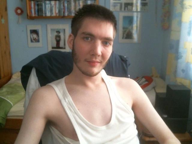 A photo of Sweetbody23-hot with short brown hair, a light beard, and a white tank top showing sideboob, sitting on a bed, looking at the camera.