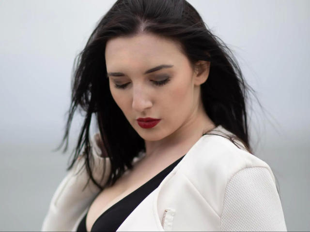 AlinaLamour-hot with black hair, red lipstick, downcast eyes, wearing a white jacket over a black top, showing her boobs. Soft, pale skin, looking cute and sexy.