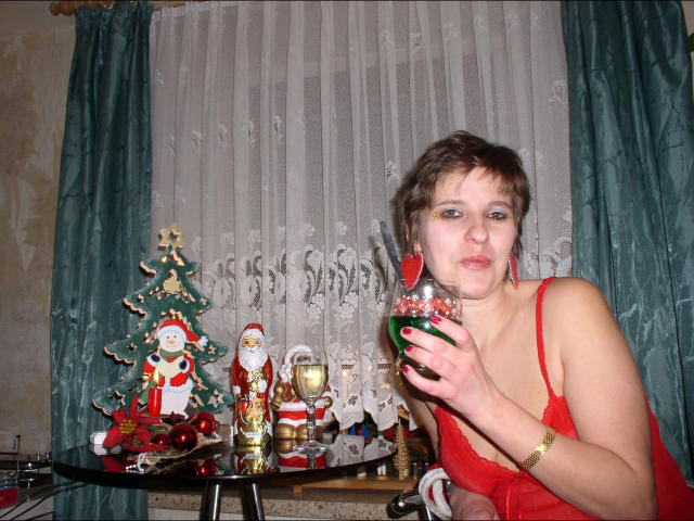 Schnuckimaus1-hot with short brown hair, red dress, and gold bracelet, sipping from a green glass, next to a Christmas tree and Santa figurines on a table.
