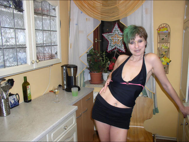 Schnuckimaus1-hot with green pixie cut, black halter top, showing small tits, black mini skirt, leaning in kitchen, bottle, glass, coffee maker, star decoration, potted plant, smile.