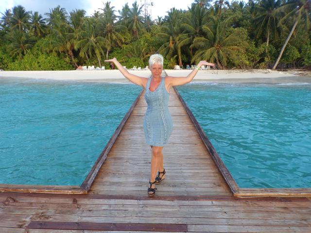 MelissaDeluxe-hot, standing on a wooden dock, arms out, wearing a blue denim dress, and black sandals, with turquoise water and palm trees in the background.