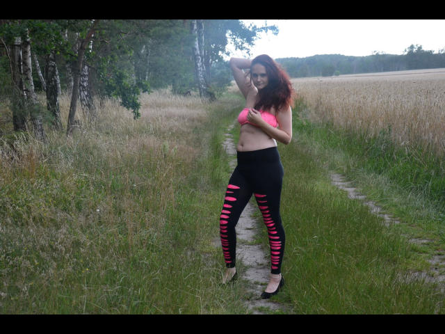 Annabell1812-hot with red hair, pink bra, and black ripped leggings standing on a grassy path, hand in hair, showing her tits, in a rural field.