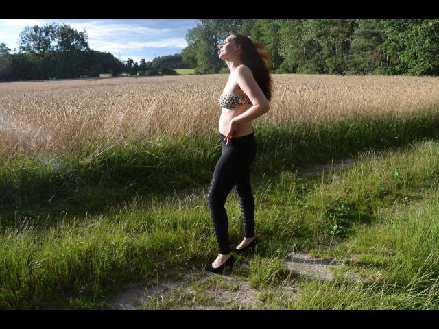 Annabell1812-hot standing in a field, wearing a leopard print bra, black skinny pants, and black heels, with long brown hair blowing in the wind.