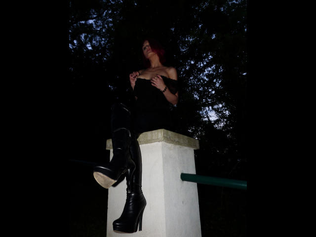 FetishQueenofPain-hot with red hair sits on a white column, wearing black thigh-high boots and an off-shoulder top, hands on her tits, in a dark, wooded area.