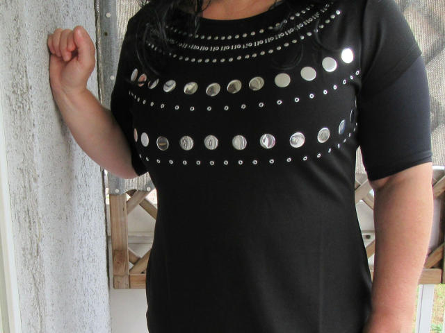 hotferkel-sex wearing a black shirt with silver studs covering their tits, standing against a textured wall, right arm raised.