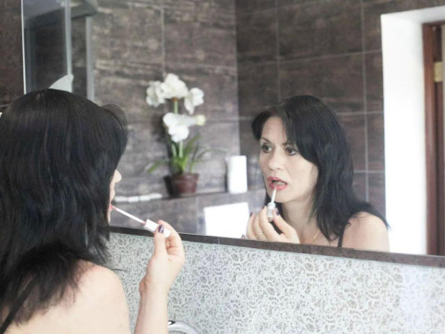 EVILENSA-hot with black hair, applying red lipstick in a bathroom mirror, holding a lipstick tube, looking at herself, reflection visible, dark brown tiled walls, white orchid in background.