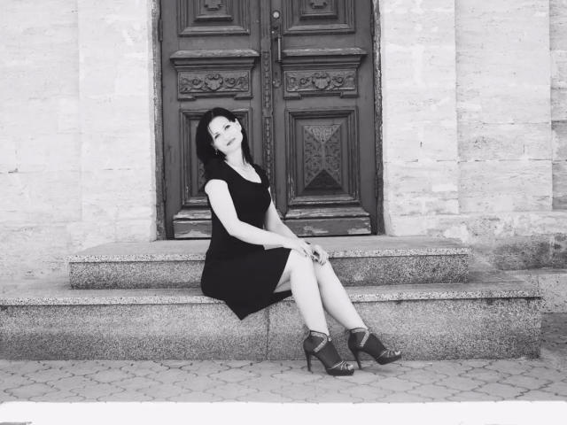 EVILENSA-hot with dark hair, black dress, and high heels, sitting on stone steps, legs crossed, smiling, hands on knees, black door behind her, black and white photo.