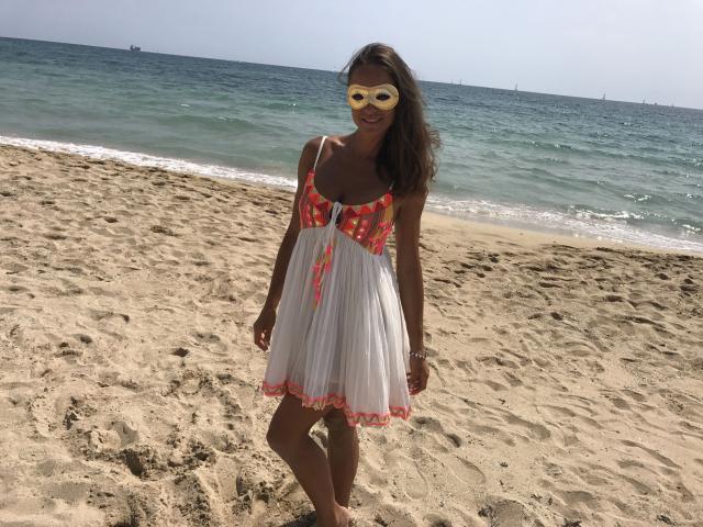 LorenaBella, con un aspecto atractivo y una máscara amarilla, un vestido blanco con estampados florales rojos, de pie en una playa arenosa, con un océano azul y un cielo despejado. Está sonriendo, tiene el cabello castaño y lleva una pulsera..