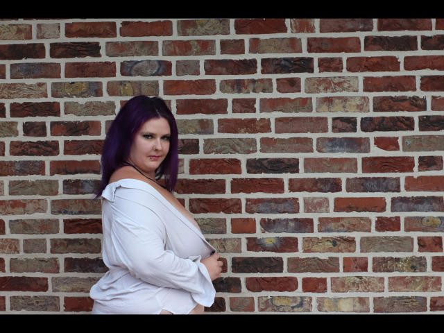 Joyckyra91-hot with purple hair, white shirt, exposing her tits, standing against a brick wall, looking at the camera with a slight smile. Her shirt is unbuttoned.