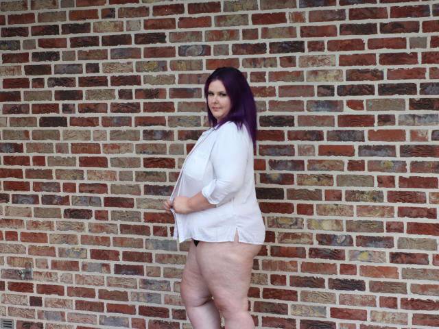 Joyckyra91-hot stands against a brick wall, wearing a white shirt, showing her big ass and thick thighs, with purple hair, looking to the side with a slight smile.