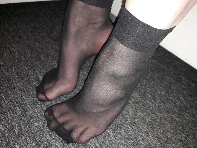 JessicaGold-hot's feet in sheer black stockings, showing her toes through the fabric, standing on a gray carpet. Her legs are slightly crossed, highlighting the texture.