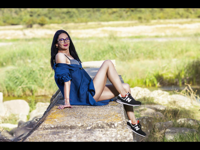SinaRauch-hot, black-haired, wearing glasses, blue off-shoulder dress, showing tits, sitting on concrete, legs up, black sneakers, green grass, rocks, sunny, attractive, sexy.