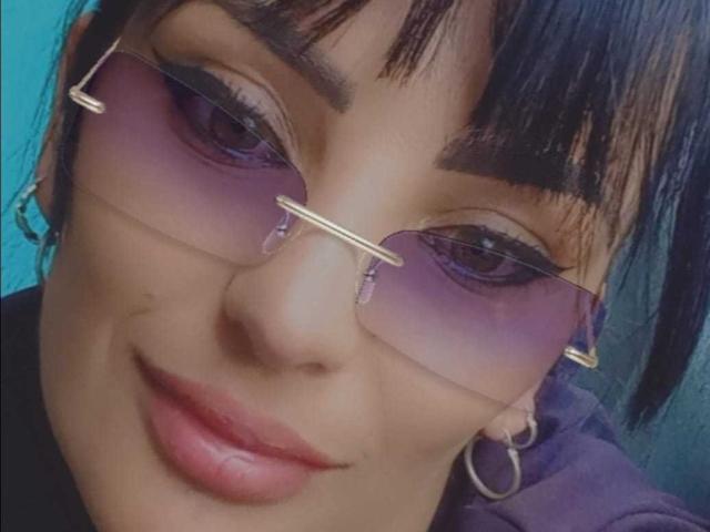 Una ragazza di nome Crazy Rouse, con occhiali di colore viola, sopracciglia nere e un sorriso appena accennato, indossa una felpa viola e orecchini a cerchio. Le sue labbra sono leggermente dischiuse. Ha i capelli scuri..