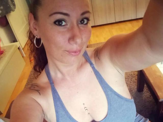 Vivien-Molinas, met kleine borsten, een blauw tanktopje, oorringen en een piercing in haar borst, maakt een selfie. Ze heeft een tatoeage op haar rechter schouder en bruin haar..