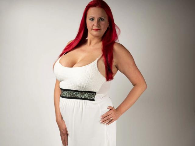 Goddess-Stone has red hair, large tits, and a white dress with a black, glittery belt. She's standing with one hand on her hip, smiling.