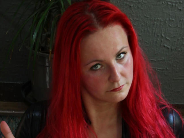 Goddess-Stone with bright red hair, green eyes, and a slight smirk, wearing a black leather jacket, has fair skin and a subtle blush on her cheeks.