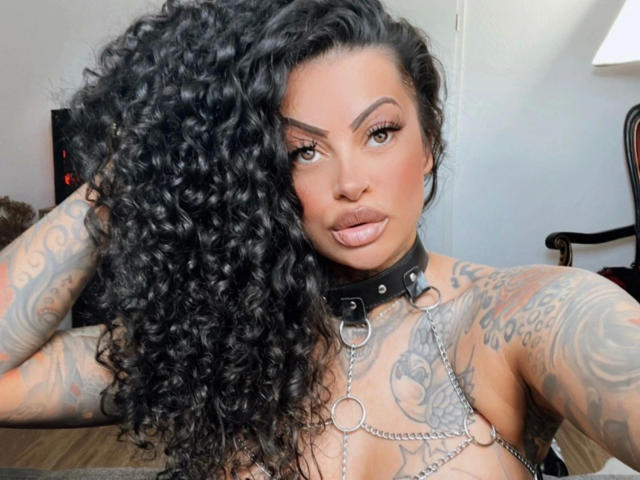 Leyla-del-Amor with huge curly black hair, tattoos on arms and chest, black leather collar with metal rings, pouty pink lips, looking at camera, holding hair with one hand.