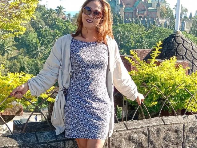xUrDestinyTS69x-hot with brown hair, wearing a blue patterned dress, white cardigan, and sunglasses, smiling, standing by yellow bushes, holding plant stems, sunny day, green background.